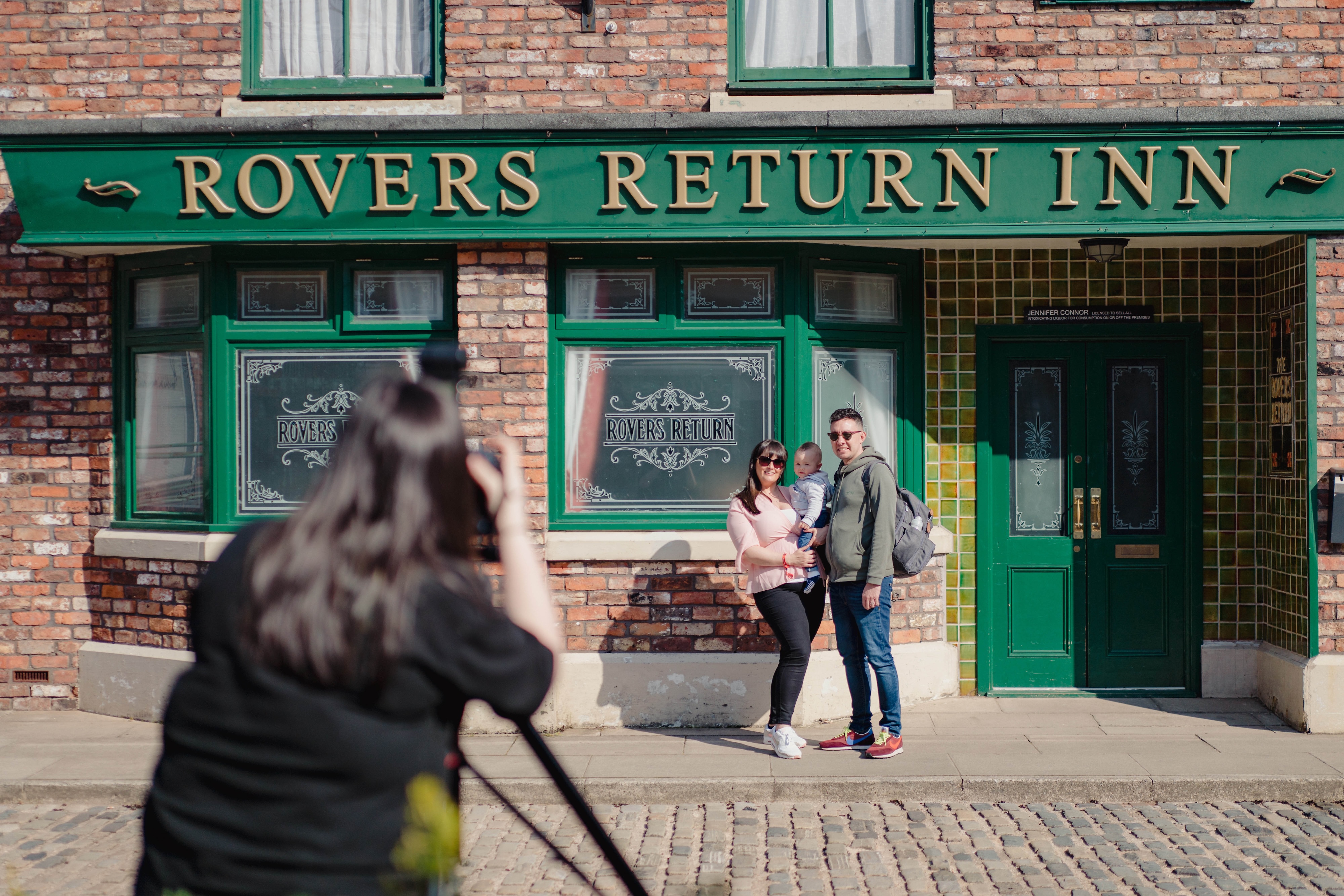 A couple with their toddler posing for a photo in front of the Rovers Return Inn at the Coronation Street Experience in Manchester