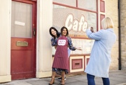 Two visitors posing for a photo in front of Roy’s Rolls Café at the Coronation Street Experience, with one wearing an apron