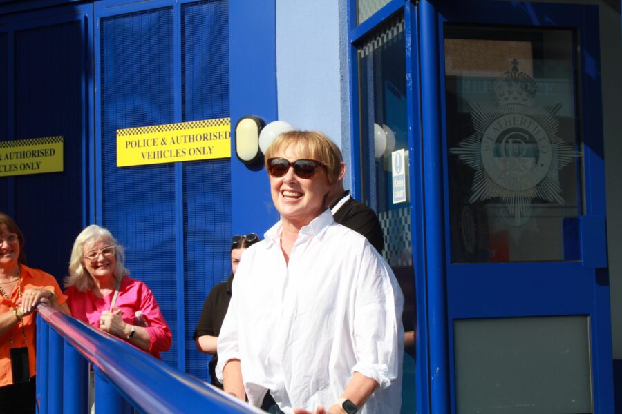 Sally Dynevor, wearing sunglasses, surprising a group of excited guests on a sunny day outside the Weatherfield police station.